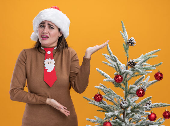 圣诞节年轻漂亮的女孩戴着圣诞帽 打着领带 站在圣诞树旁 双手分开放在橙色的背景上领带树未租赁