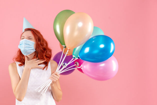 病毒正面图是粉红色墙上戴着消毒面具手持彩色气球的年轻女子生日无菌圣诞节