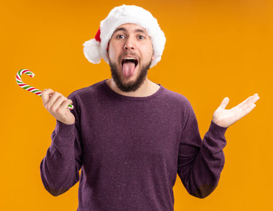 舌头身穿紫色毛衣 戴圣诞帽的年轻人手持糖果手杖 看着伸出舌头的相机 站在橙色的背景上 快乐而愉快年轻毛衣圣诞老人