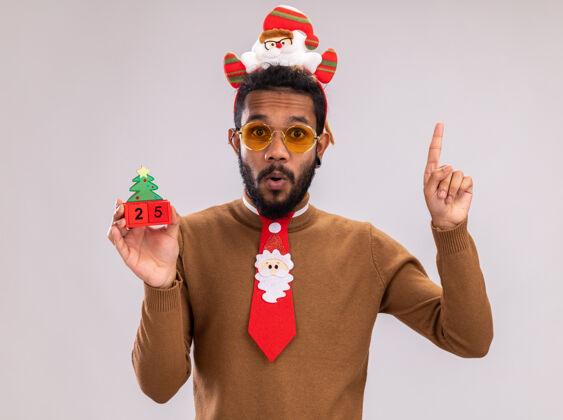 惊讶一个非洲裔美国人 穿着棕色毛衣 头上戴着圣诞老人的戒指 打着有趣的红色领带 手里拿着日期为25的玩具立方体 惊讶地看着相机 食指站在白色背景上手指毛衣非洲