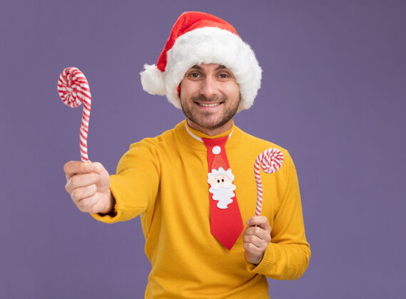 微笑微笑着的年轻白人男子戴着圣诞帽 打着圣诞领带 手里拿着圣诞甜甜的手杖 一根手杖伸向镜头 看着镜头孤立地放在紫色背景上 留着复印空间领带手杖抱着