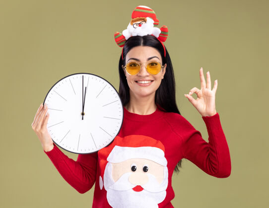 时钟微笑着的年轻白人女孩戴着圣诞老人的头带和毛衣 戴着眼镜 拿着时钟 看着相机 在橄榄绿的背景上做着“ok”的手势女孩微笑圣诞老人