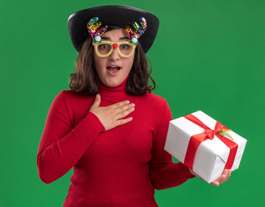女孩穿着红色毛衣的小女孩戴着滑稽的眼镜 戴着黑色的帽子 拿着礼物看着相机 惊讶而快乐地站在绿色的背景上拿着年轻礼物