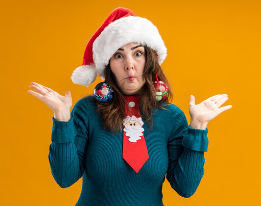 打开惊讶的成年白人妇女戴着圣诞帽 打着圣诞领带 耳朵上戴着玻璃球饰物 做着鱼嘴 在橙色背景上保持双手张开 并留有复制空间领带玻璃装饰品