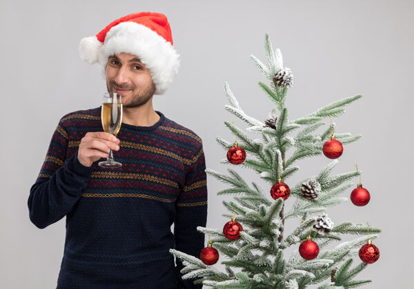 抱着高兴的年轻白人男子戴着圣诞帽站在圣诞树旁拿着一杯香槟看着隔离在白色背景下的相机高兴圣诞树年轻人