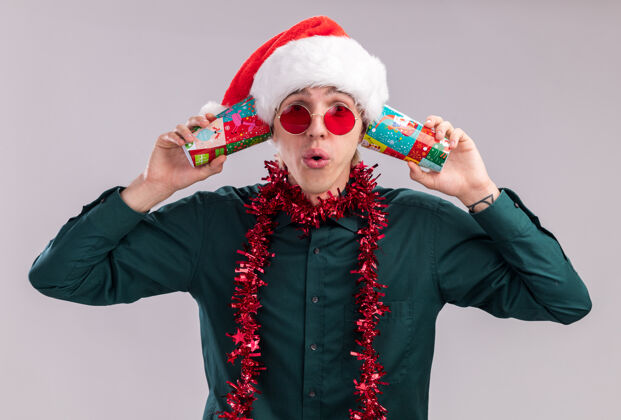圣诞节令人印象深刻的年轻金发男子戴着圣诞帽 戴着眼镜 脖子上戴着金属丝花环 耳边拿着塑料圣诞杯 听着秘密 看着隔离在白色背景上的相机塑料花环圣诞老人
