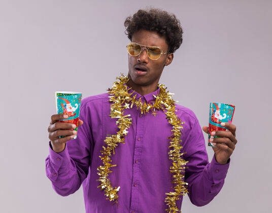 年轻恼怒的年轻美国黑人男子戴着眼镜 脖子上戴着金属丝花环 手里拿着塑料圣诞杯 看着其中一个孤立的白色背景杯子塑料花环