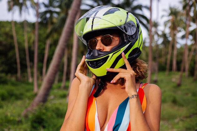乐趣在棕榈树下热带田野的丛林中 穿着黄绿色摩托车头盔和彩色浅夏装的美丽骑手妇女的肖像滑板车旅游骑自行车