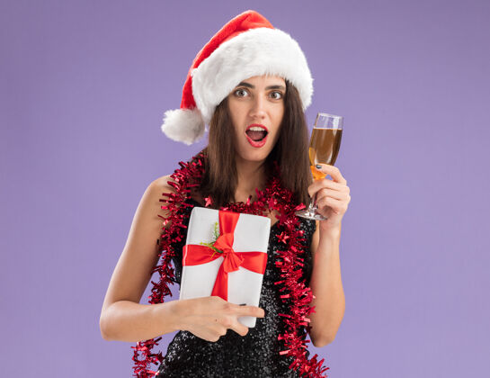 年轻惊讶的年轻漂亮女孩戴着圣诞帽 脖子上戴着花环 手里拿着一个礼盒 紫色背景上隔着一杯香槟玻璃杯女孩盒子