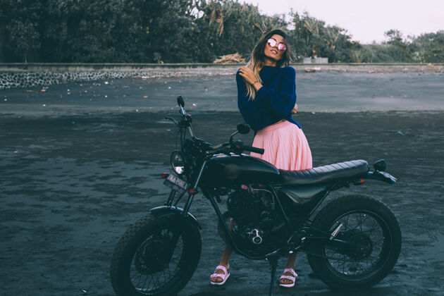 漂亮戴着太阳镜 穿着蓝色毛衣和粉色裙子的时髦女人的时尚肖像 站在黑色沙滩上的黑色自行车旁汽车骑自行车女孩自行车