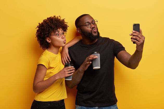 非洲搞笑的黑皮肤情侣对着手机摄像头撅起嘴唇 自拍写真 从一次性杯子里喝咖啡去 穿黑黄相间的t恤关系饮料饮料