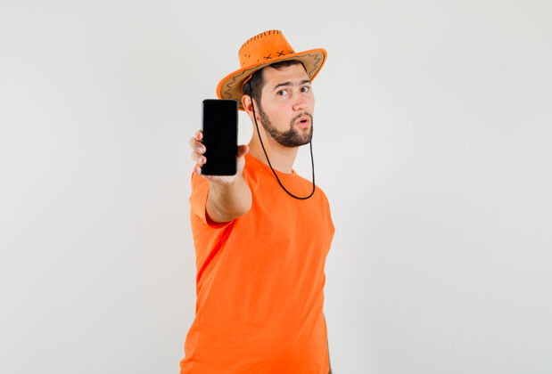 年轻年轻人展示手机在橙色t恤 帽子 前视图男人男人成人