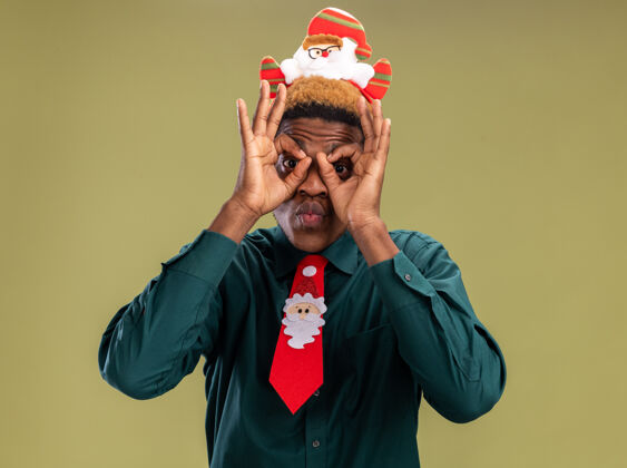手势快乐的非洲裔美国人 带着滑稽的圣诞老人 站在绿色的背景上 用手指做着双目手势看着相机美国手指搞笑