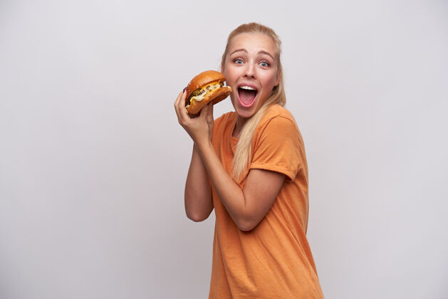食物摄影棚照片中 年轻的蓝眼睛兴奋的金发女郎身着橙色t恤 双手举着大汉堡 睁大眼睛 张大嘴巴看着相机 与白色背景隔绝饮食年轻美味
