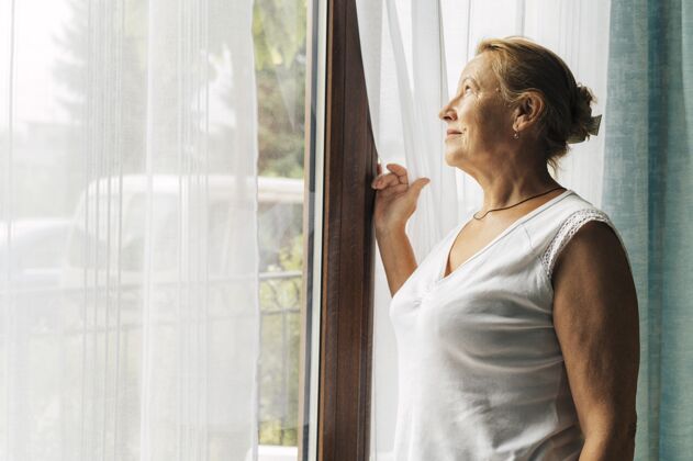 隔离大流行期间家里的老太太透过窗户看距离防护社会距离
