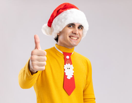 圣诞老人快乐的年轻人穿着黄色高领毛衣 戴着圣诞老人帽 打着滑稽的领带 面带微笑地看着镜头 站在白色背景上竖起大拇指年轻高领毛衣帽子