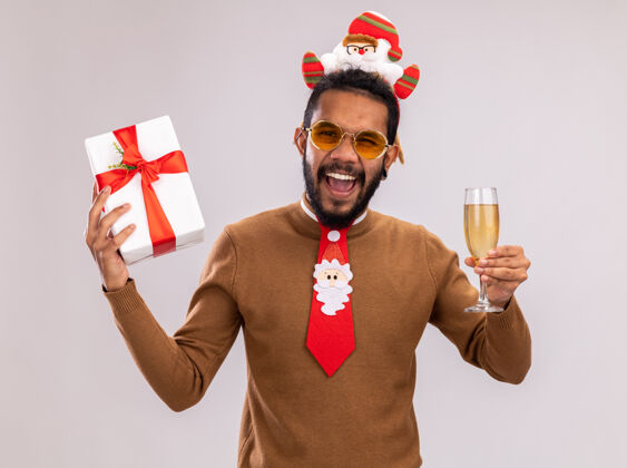 领带一个身穿棕色毛衣 头戴圣诞老人戒指 打着有趣的红色领带 手里拿着礼物和一杯香槟的非裔美国人站在白色背景下兴奋而快乐地尖叫着礼物尖叫毛衣
