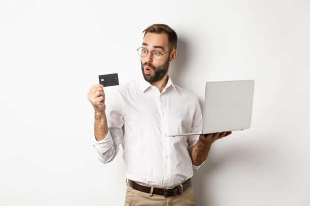 经理网上购物惊讶的商人拿着笔记本电脑 看着印象深刻的信用卡 站着手势支付电子商务