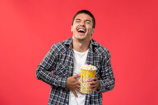 容器正面图：年轻男子拿着爆米花包 笑着站在浅红的墙上看电影年轻男性包装电影院