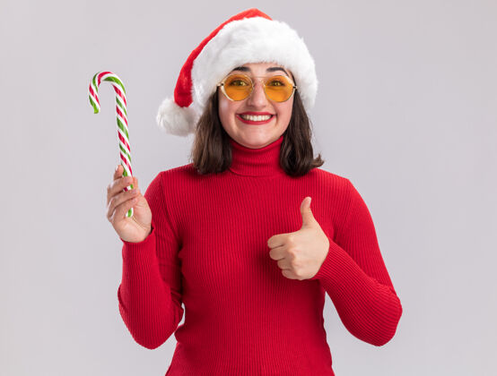 站着快乐的年轻女孩穿着红色毛衣 戴着圣诞帽 戴着眼镜 手持糖果手杖 微笑着看着镜头 竖起大拇指站在白色背景上圣诞老人微笑手杖