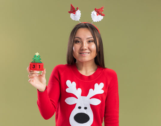 亚洲人笑容满面的年轻亚洲女孩戴着圣诞发箍 拿着橄榄绿背景上孤立的圣诞玩具抱着圣诞节篮球