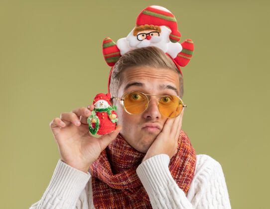 焦虑焦虑的年轻帅哥戴着圣诞老人的头带和围巾看着相机拿着雪人圣诞饰品手放在脸上橄榄绿的背景隔离相机快乐抱