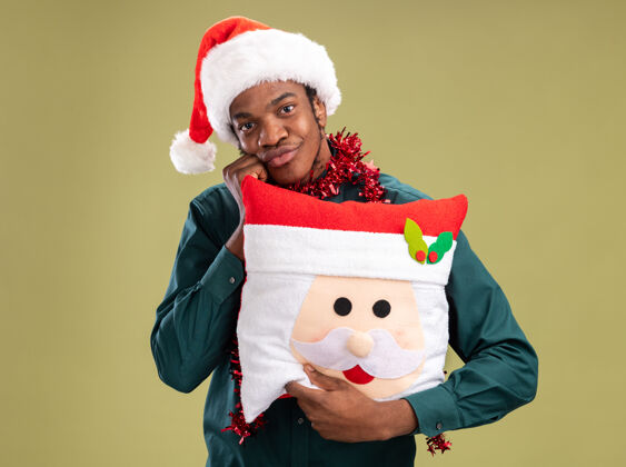 抱着快乐的非洲裔美国人戴着圣诞帽 戴着花环 抱着圣诞枕头 面带微笑地看着相机 站在绿色的背景上圣诞老人圣诞节帽子