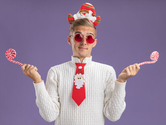 看着高兴的年轻帅哥戴着圣诞老人的头带和领带戴着眼镜拿着圣诞糖果手杖看着隔离在紫色背景上的相机领带圣诞老人帅哥