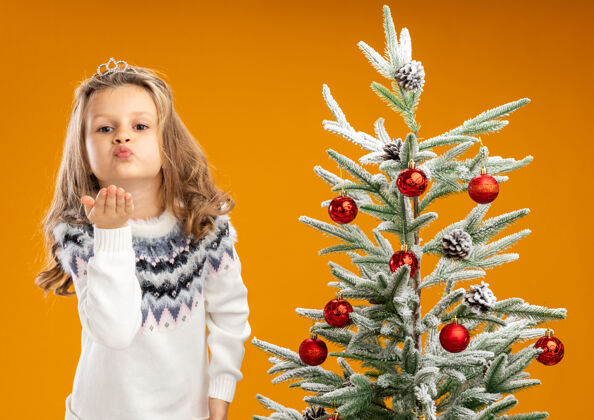 圣诞树可爱的小女孩站在圣诞树旁 戴着头饰 脖子上戴着花环 在橙色背景上显示出孤立的亲吻手势花环小高兴