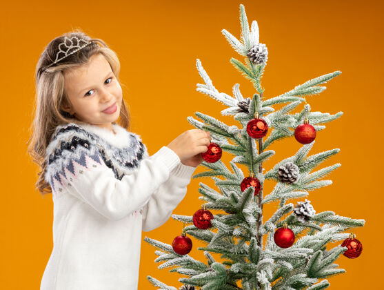 挂起来高兴的小女孩站在圣诞树旁边戴着头饰 脖子上戴着花环 把玩具挂在树上孤立的橙色背景上圣诞树脖子快乐