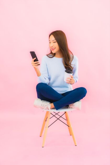 站立肖像美丽的亚洲年轻女子在粉色隔离墙上使用智能手机手机休闲时尚