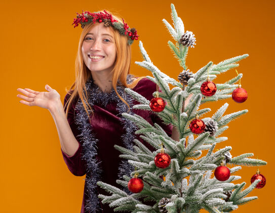 伸展微笑着站在圣诞树后面的年轻漂亮的女孩穿着红色的裙子 脖子上戴着花环 手被隔离在橙色的背景上女孩微笑手