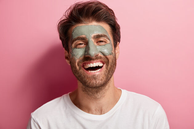 面膜快乐快乐的男人脸上有泥面膜 享受水疗 笑容灿烂 精神高昂 关心美丽牙齿干燥护肤