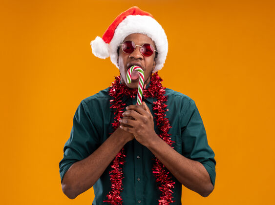 眼镜戴着圣诞帽 戴着花环 戴着眼镜 拿着糖果手杖的非裔美国人站在橙色背景下 兴高采烈地微笑着看着摄像机非洲人微笑花环