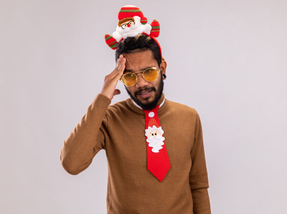 非洲一个非洲裔美国人 穿着棕色毛衣 头上戴着圣诞老人的戒指 戴着一条有趣的红色领带 站在白色的背景下 他看起来很不舒服 头痛边缘触摸美国