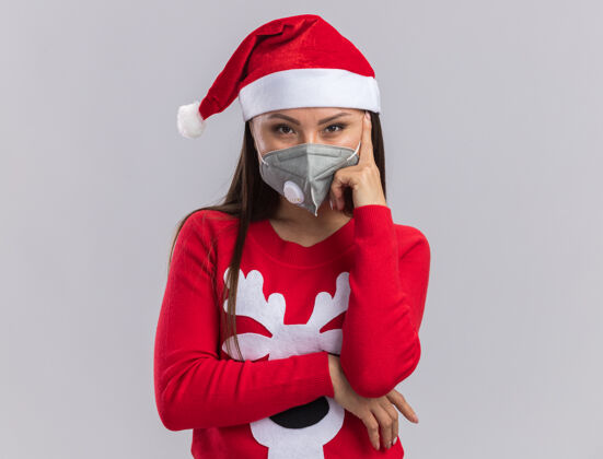女孩自信的年轻亚洲女孩戴着圣诞帽 穿着毛衣 戴着医用口罩 手指贴在脸颊上 白色背景 与世隔绝面具医学脸颊