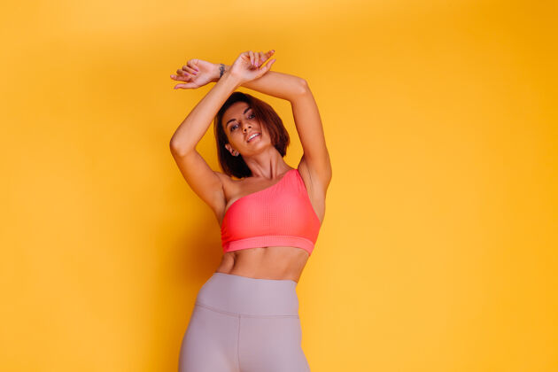 有力年轻健壮的女人穿着运动服 时髦的上衣和紧身裤 对着黄色的墙壁摆姿势运动身材锻炼