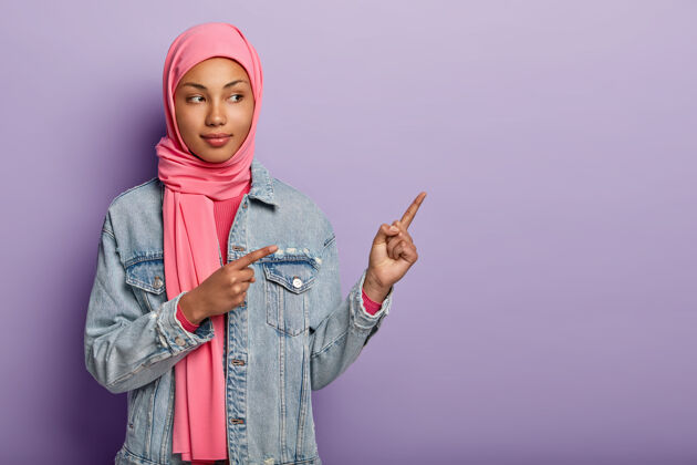 食指一张自信的阿拉伯女士戴着传统粉色头巾的照片 告诉你正确的方向 用两个前指指着空白处 邀请你去那里 促销员复制空间和我一起看看面纱女士侧边