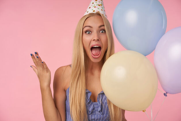 女士快乐迷人的年轻女子 金色长发 随意的发型 在收到令人敬畏的礼物时表现出愉快的反应 站在粉色背景下 摆出五颜六色的气球气球正面20多岁
