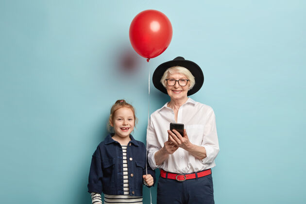 孩子微笑的奶奶戴着黑色时髦的帽子 穿着白色优雅的衬衫和正式的裤子 手里拿着手机 懂得如何用好现代的小玩意儿 为手持红色气球的小孩庆祝生日高兴小玩意短信