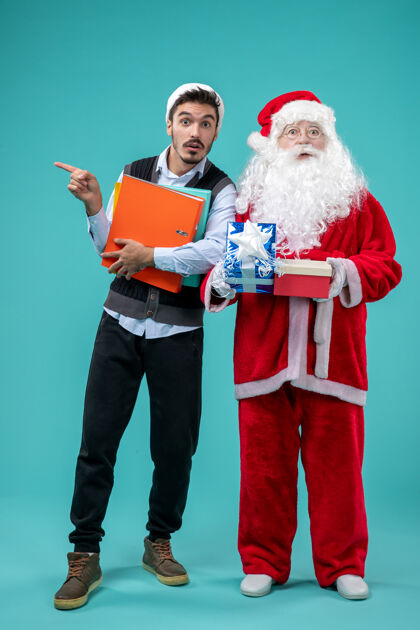 假期圣诞老人与年轻男性和礼物在蓝色墙上的正面视图礼物节日音乐家