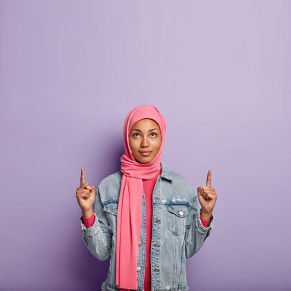 促销严肃的年轻妇女的照片 用两个前指向上指着 戴着粉色围巾 穿着粉色牛仔夹克 隔着紫色的墙人 广告和促销广告表情个人