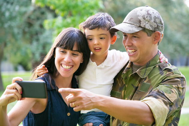 拍摄快乐兴奋的军人 他的妻子和小儿子在城市公园里用手机自拍正面视图家庭团聚或回家的概念统一爸爸男人