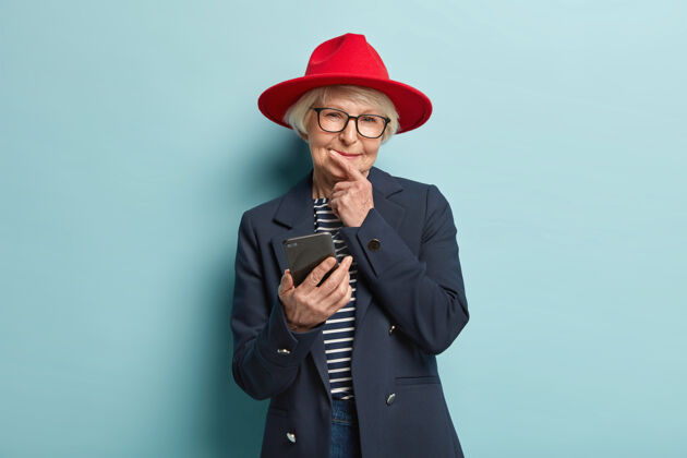 电话睿智体贴高兴的老皱纹妇女托着下巴 阅读通知 连接无线互联网 戴着红色头饰和时尚外套 在电子邮件上获得折扣人 年龄 智慧智慧老年人应用程序