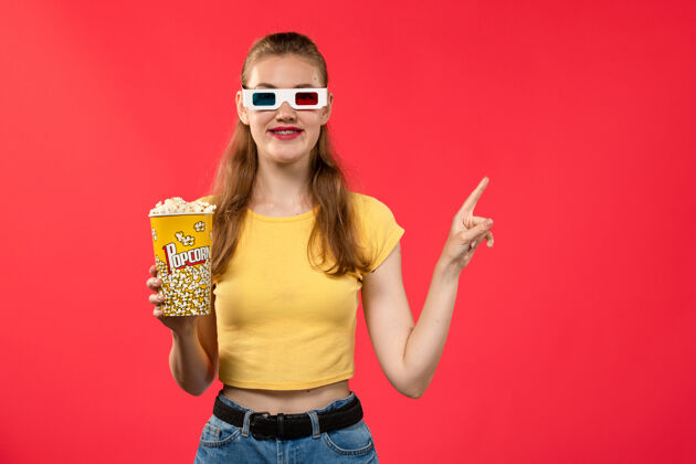 漂亮前视图年轻的女性在电影院拿着爆米花包在d太阳镜在浅红色的墙上电影院有趣的电影成人电影院年轻女性