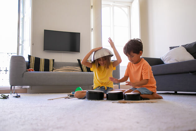 儿子金发碧眼的女孩头上抱着碗和朋友玩耍快乐的小男孩敲盘子两个快乐的孩子坐在地板上 在客厅里一起玩童年 假日和家的概念地毯年轻孩子