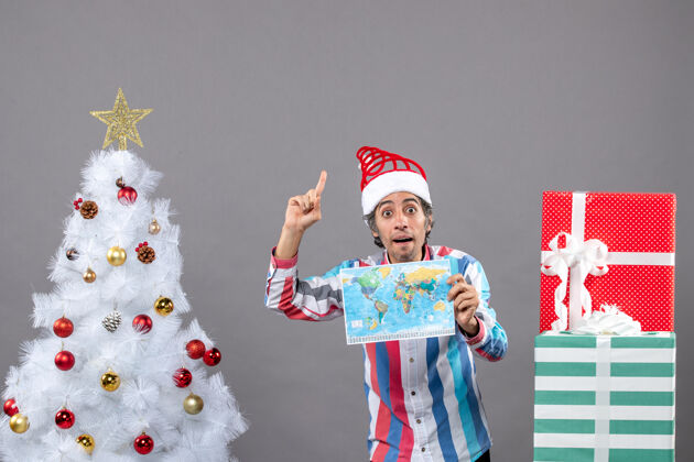 灰色前视图惊讶的男子戴着螺旋弹簧圣诞帽望着高高的世界地图指着高高的地图抱着假期