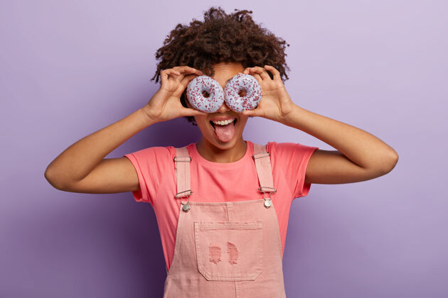 五颜六色积极的女性眼睛上拿着两个紫色的甜甜圈 伸出舌头 穿着粉色的t恤和工作服 喜欢吃甜食 玩得很开心 在紫罗兰色的墙上摆出室内姿势和我一起吃美味的甜点釉面甜甜圈甜点