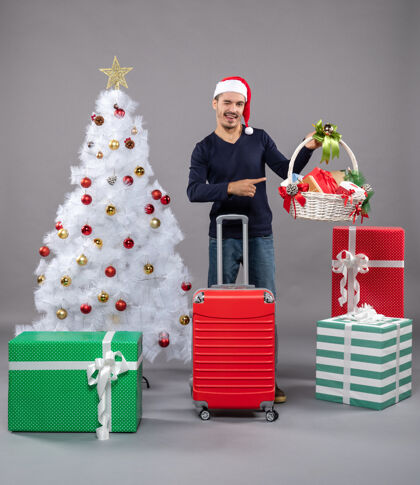 圣诞帽兴奋的年轻人拿着礼品篮站在白色圣诞树旁 灰色的圣诞树上摆着五颜六色的礼物男人盒子圣诞