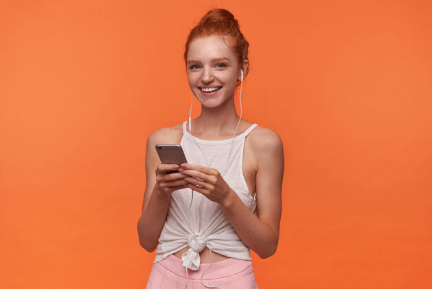 微笑迷人的年轻readhead女性的摄影棚照片 发髻在橙色背景下摆出姿势 双手举着智能手机 戴着耳机听音乐漂亮音乐年轻